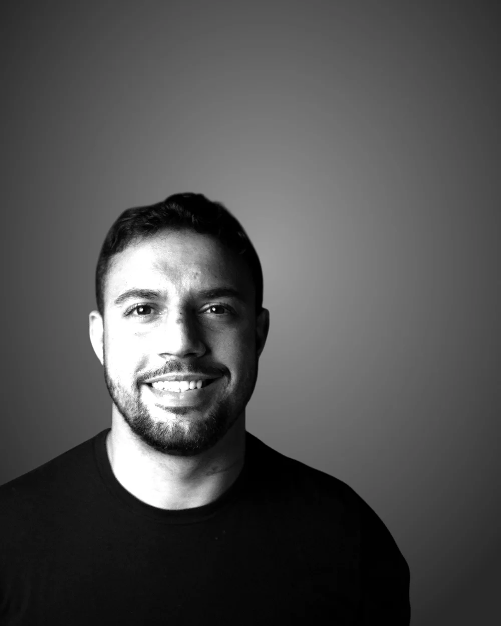 Retrato em preto e branco de Raphael Busquet, sorrindo, de camiseta escura